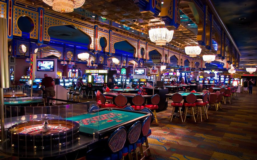 Speelzaal van een casino: roulettetafel op de voorgrond, daarachter rijen gokkasten en speeltafels.